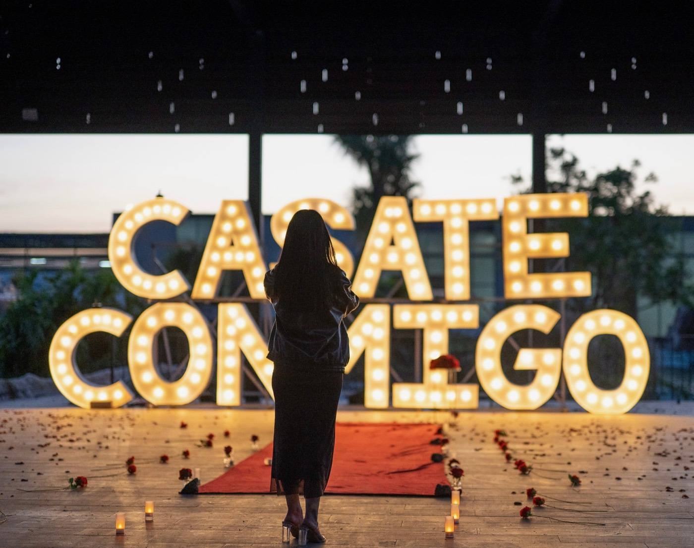 Ella frente a las letras gigantes iluminadas CÁSATE CONMIGO en un camino de pétalos