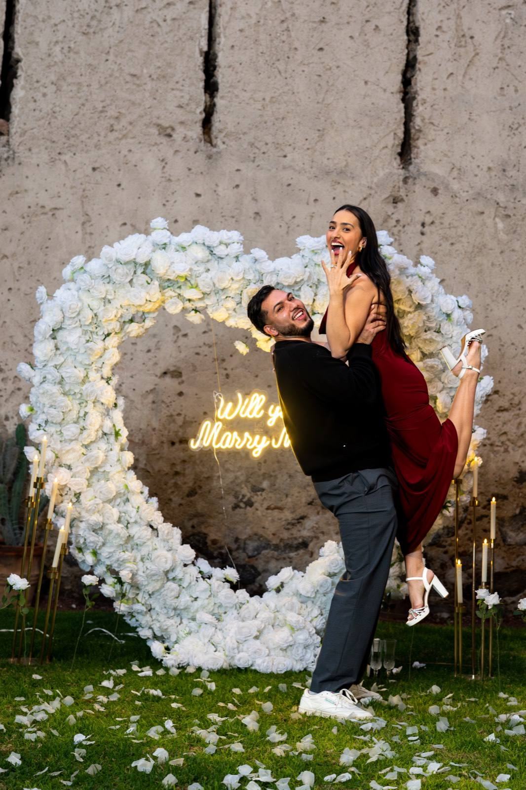 Laura y Enrique felices frente al corazón de rosas blancas de su pedida de matrimonio